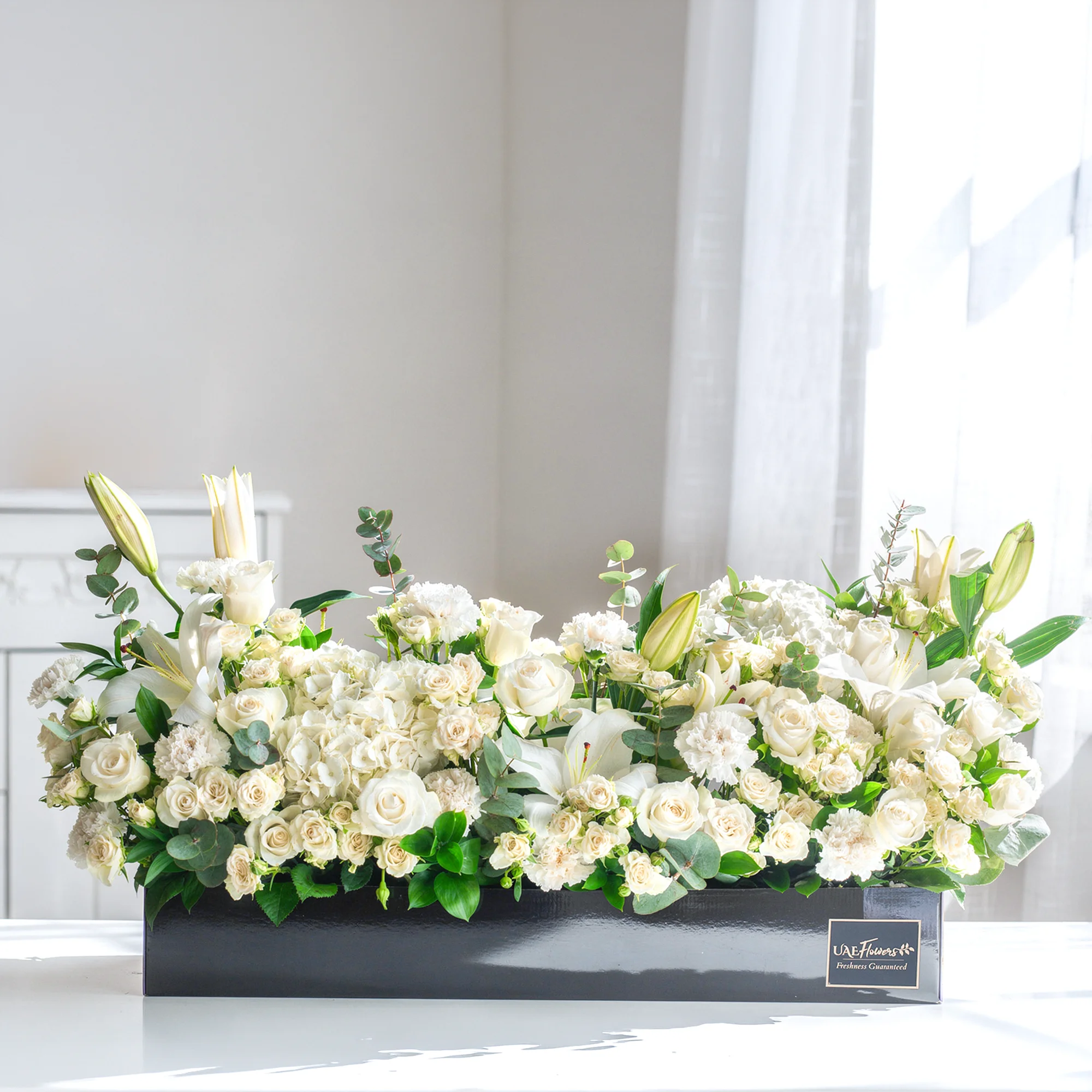 Arrangement of 5 white lilies, 20 white roses, 20 white spray roses, 10 white carnations and 2 white hydrangeas with green fillers in a long black box.