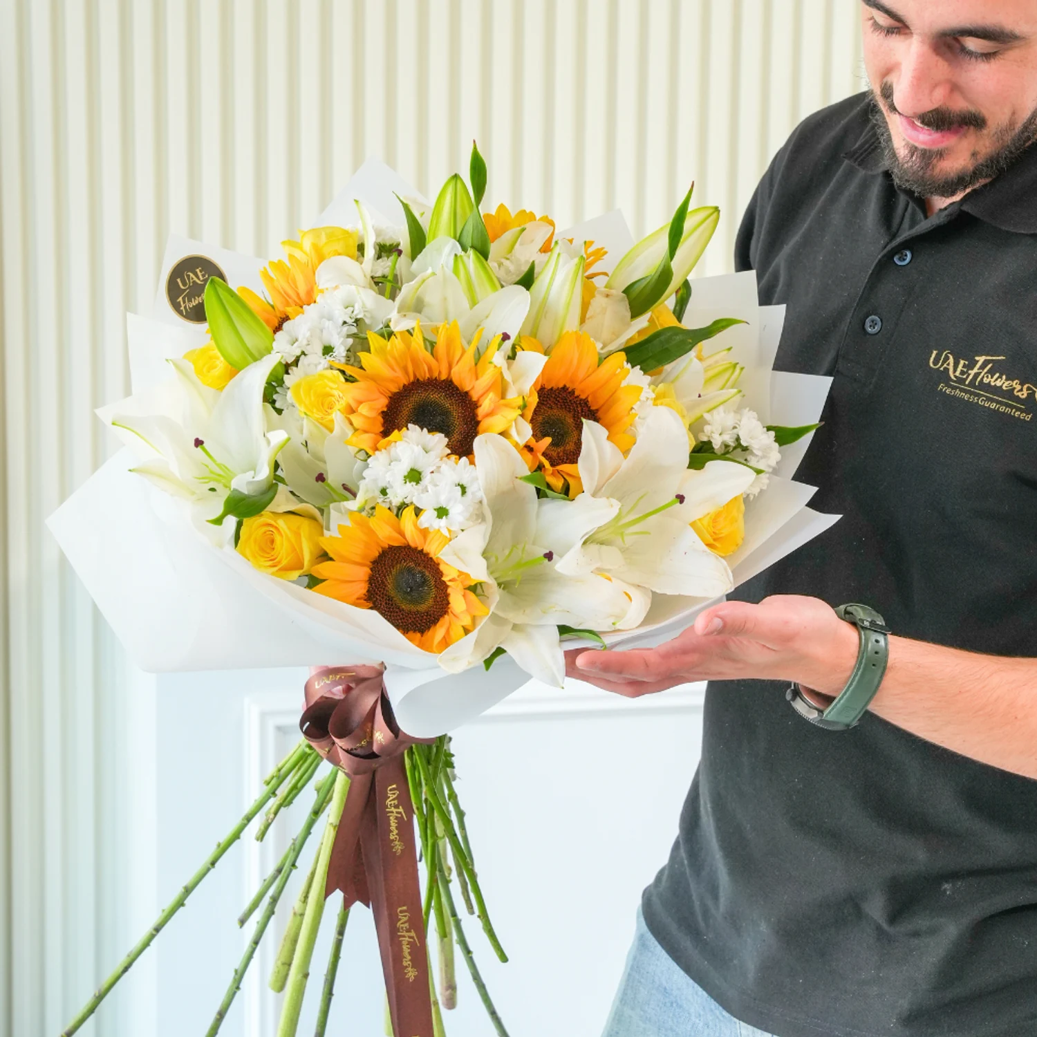 Bright bouquet of sunflowers, white lilies, yellow roses, and white filler flowers wrapped in elegant white paper with a brown satin ribbon – joyful floral gift by UAE Flowers.