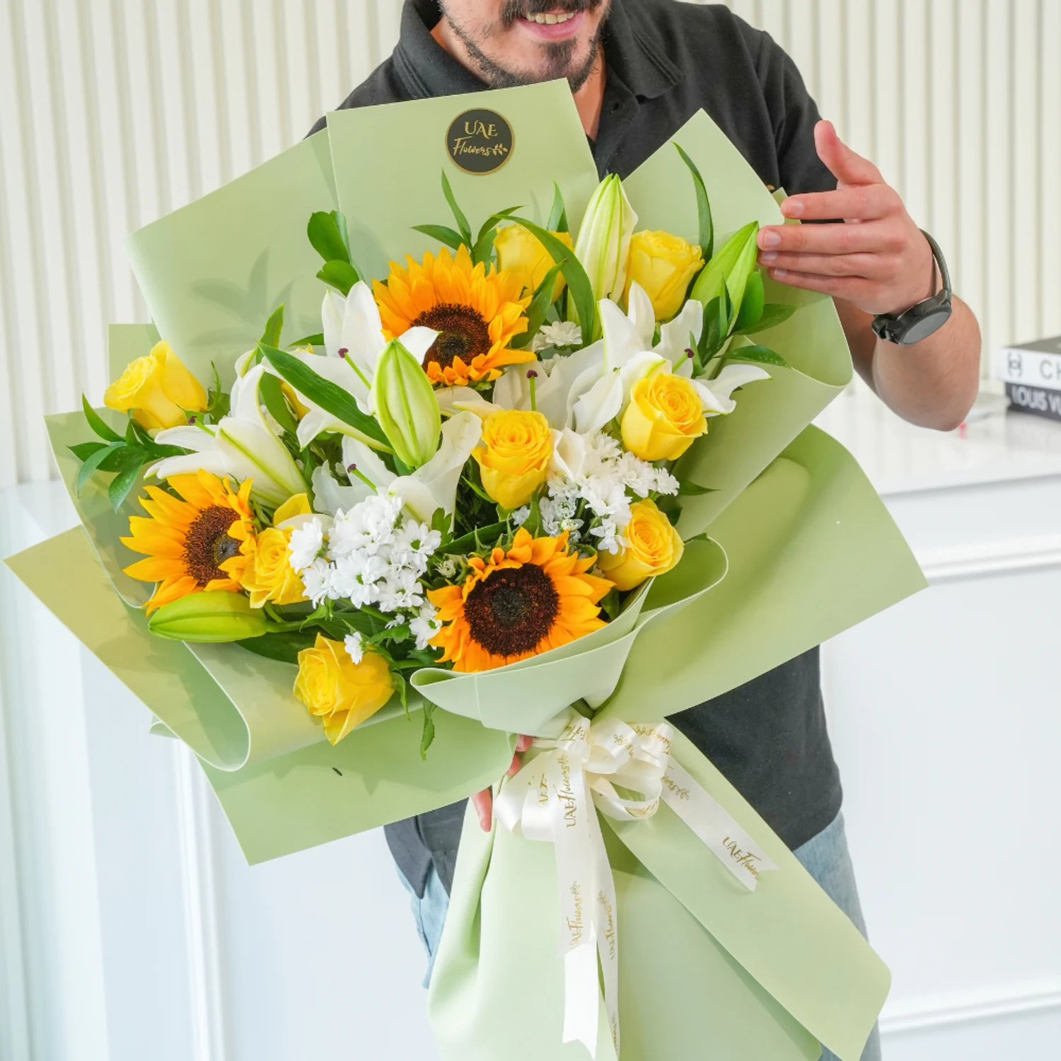 Bouquet of sunflowers, white lilies, yellow roses, and filler flowers wrapped in pastel green paper with a white satin ribbon – cheerful floral gift by UAE Flowers.