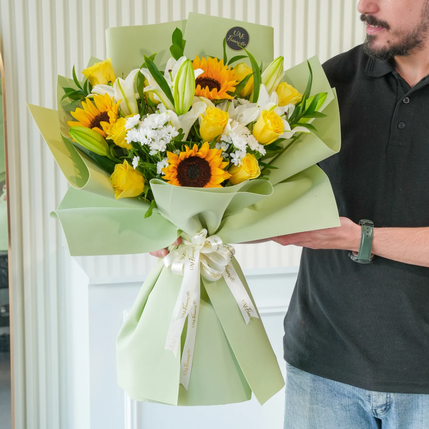 Bouquet of sunflowers, white lilies, yellow roses, and filler flowers wrapped in pastel green paper with a white satin ribbon – cheerful floral gift by UAE Flowers.