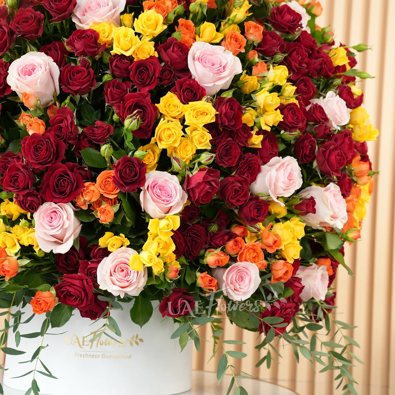 A half-round floral arrangement featuring 30 pink roses and 50 red, orange, and yellow spray roses, along with green fillers and eucalyptus leaves, presented in a white round box