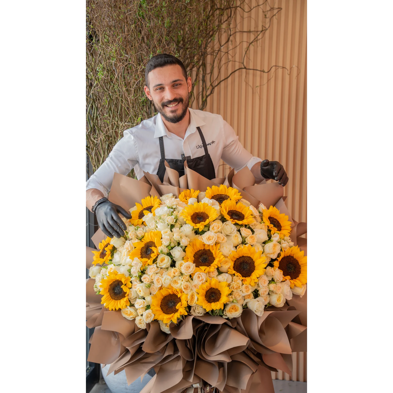 Tall luxury floral stand with 15 sunflowers, 50 spray roses, and 50 premium roses, wrapped in brown draping with satin ribbons – elegant event display by UAE Flowers.
