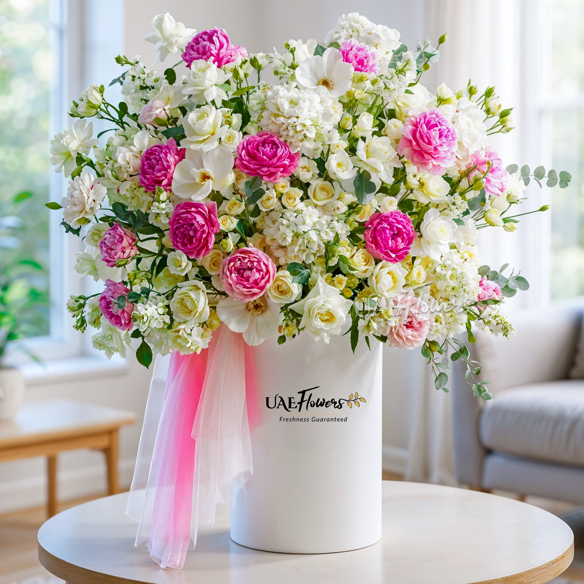 An arrangement of white hydrangeas, pink peonies, white roses, white matthiola and white spray roses with green fillers and pink & white net in a tall round box.