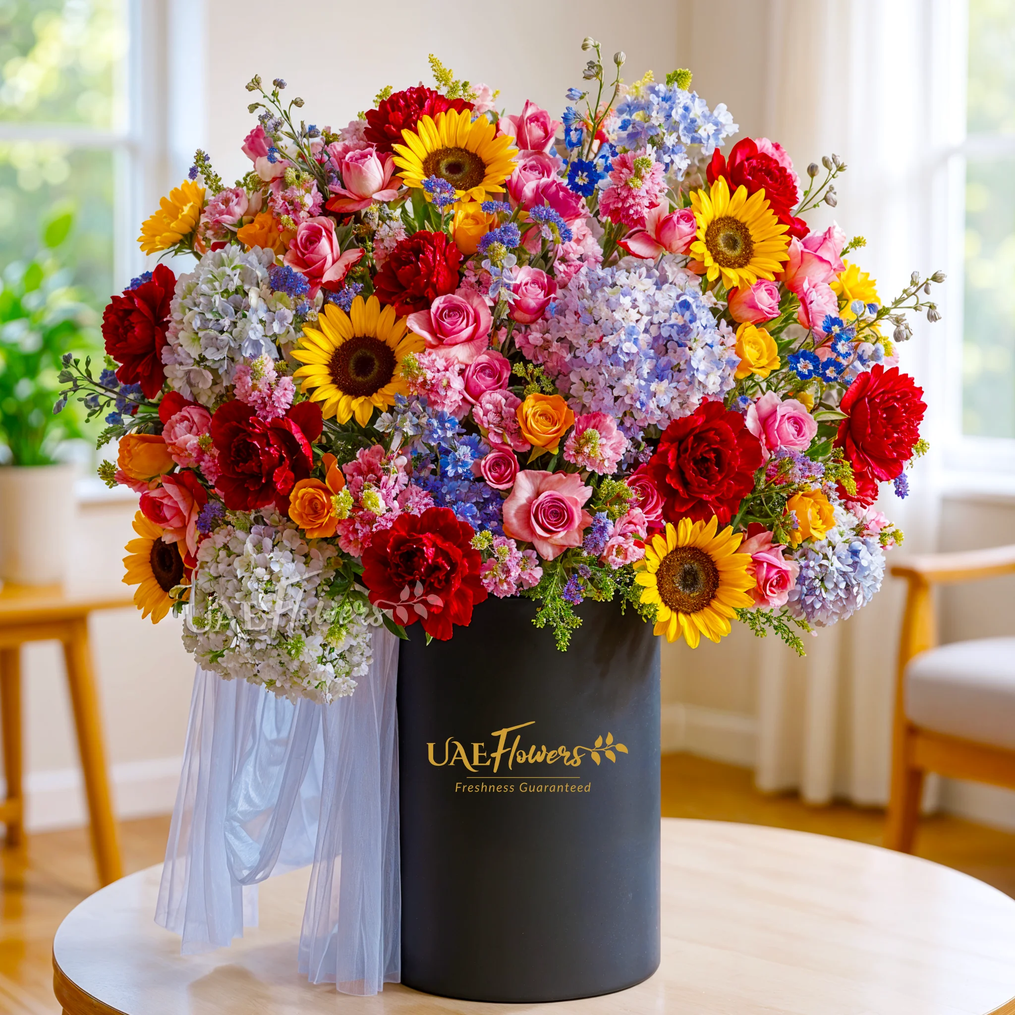 An arrangement of blue Hydrangeas, sunflowers, blue mattihola, purple lisianthus, orange roses, purple roses, red peonies with green fillers and blue net in a tall round box.